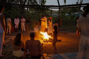 Group gathered around blazing bonfire at night in outdoor setting with tropical foliage.
