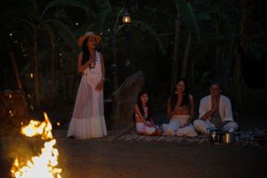 Evening gathering around campfire with performers in white dresses and audience seated on ground.