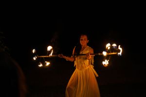 Fire performer spinning illuminated batons in a flowing white dress against dark night sky background.