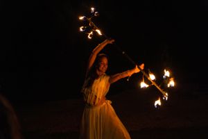 Performer in flowing dress spinning fire batons against dark night sky.