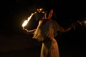 Fire dancer performing with flaming batons in white dress against night sky.
