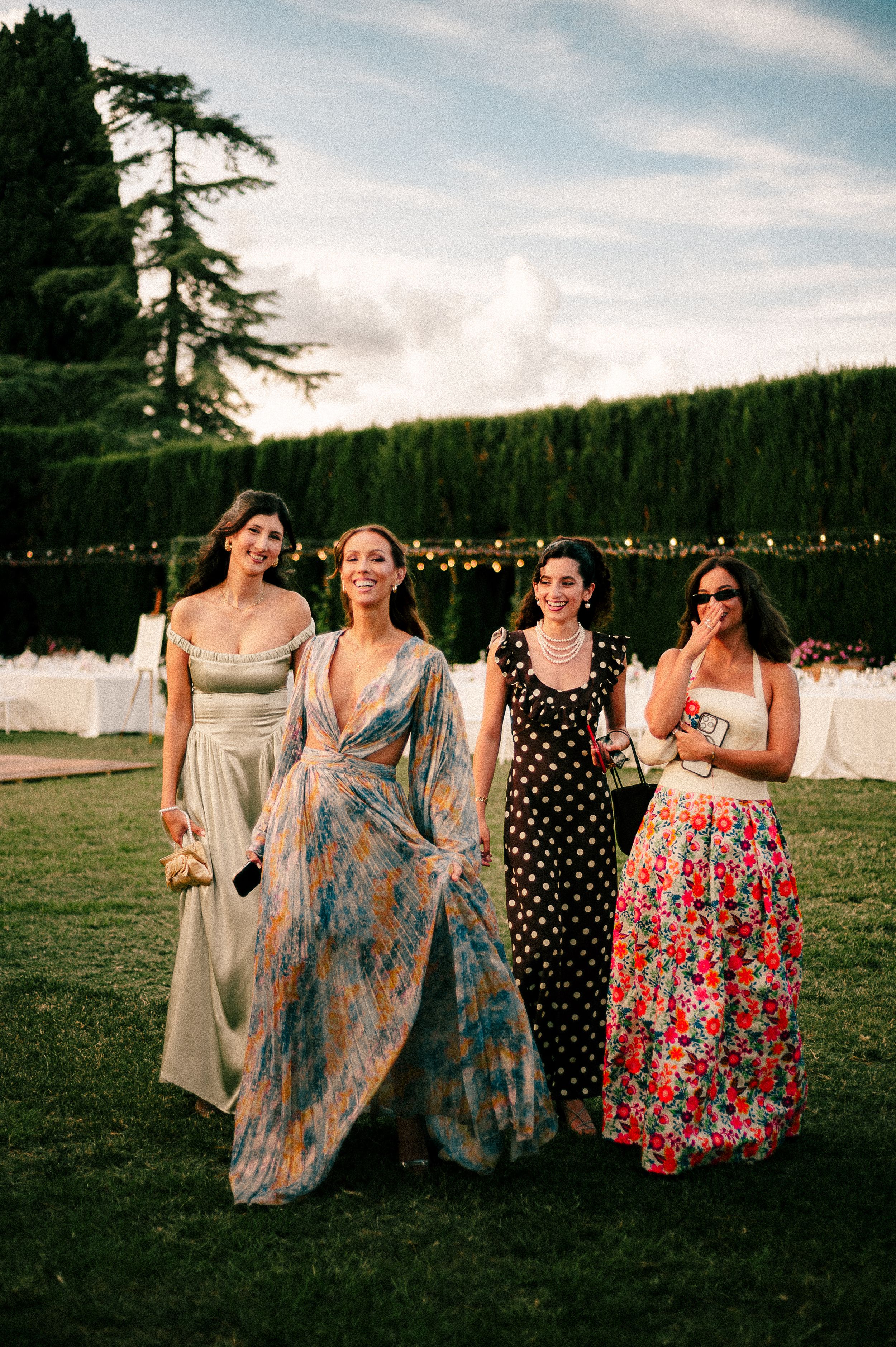Wedding guests in colorful formal dresses stand on a lawn with string lights and hedges.