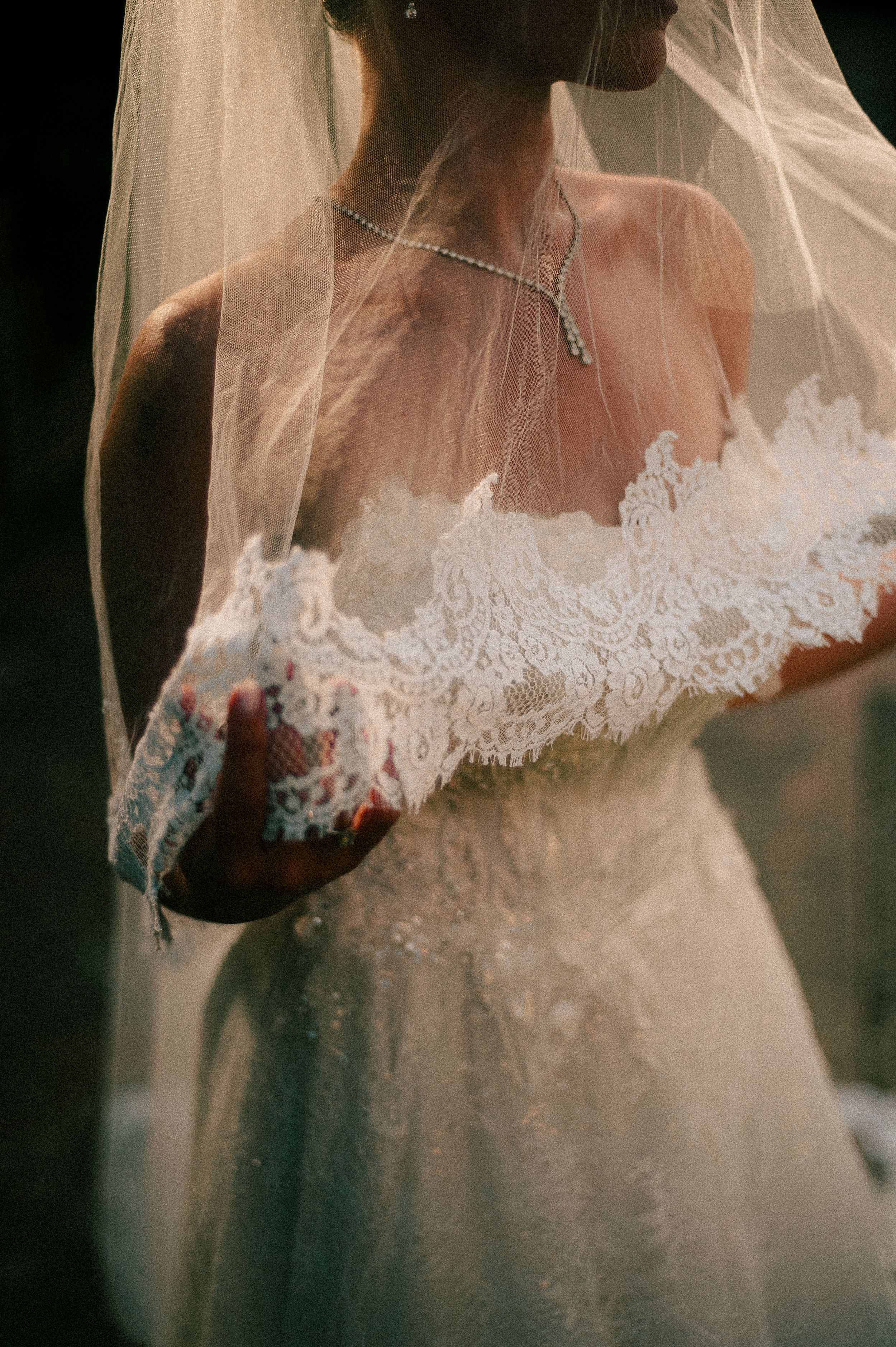 Bride wearing delicate lace wedding dress with flowing veil and necklace in soft romantic lighting.