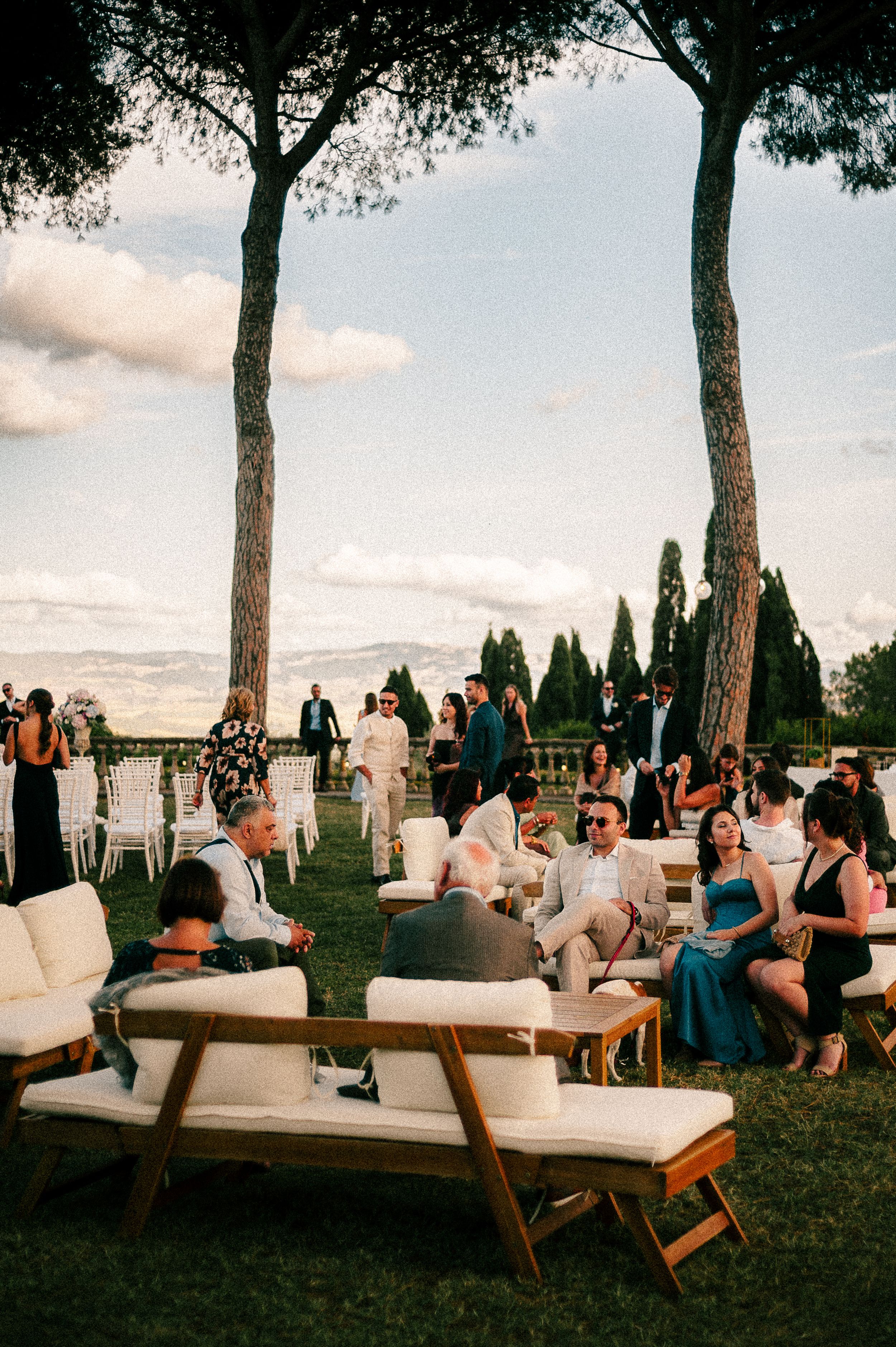 Wedding guests relax on stylish outdoor lounge furniture under tall trees at sunset.
