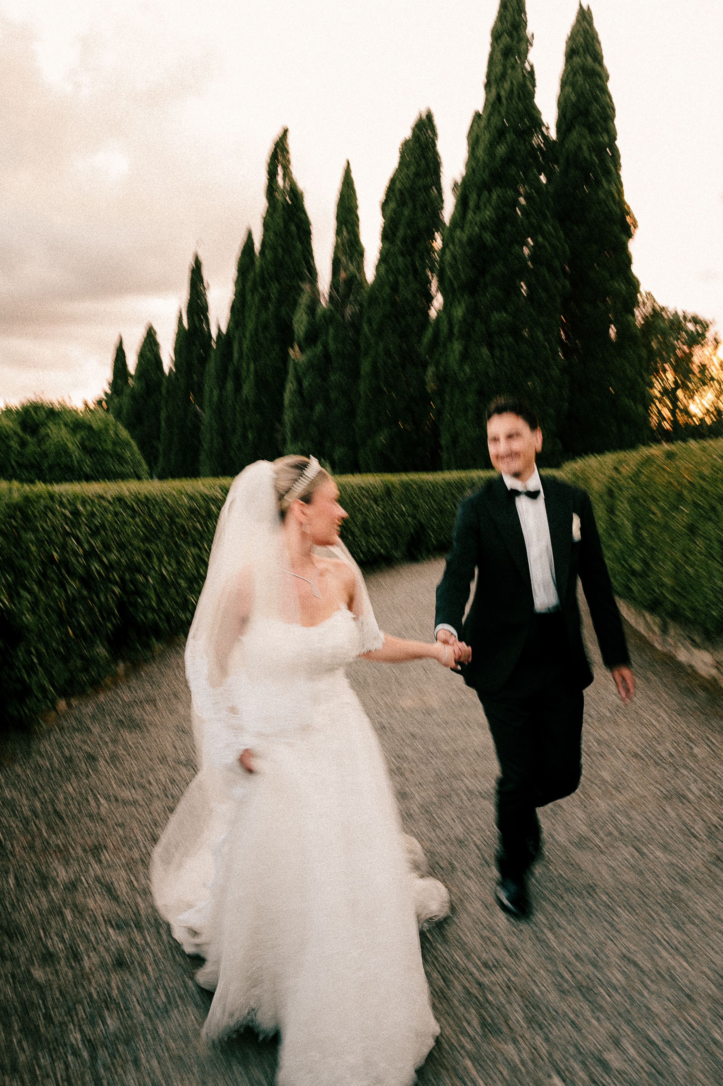 A wedding couple walks along a cypress tree-lined path in romantic evening light.