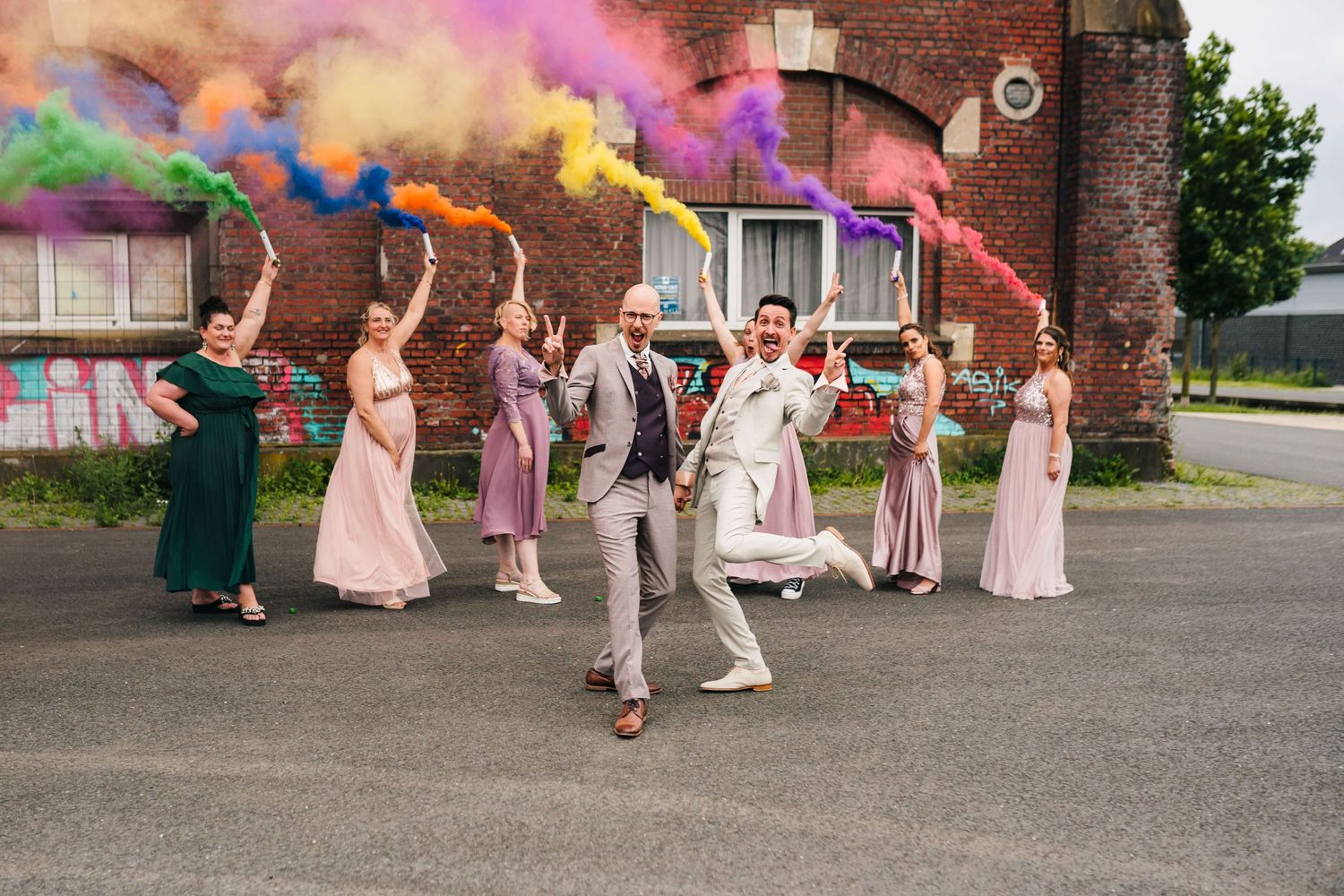 Wedding party celebrates with colorful smoke bombs outside a brick building.