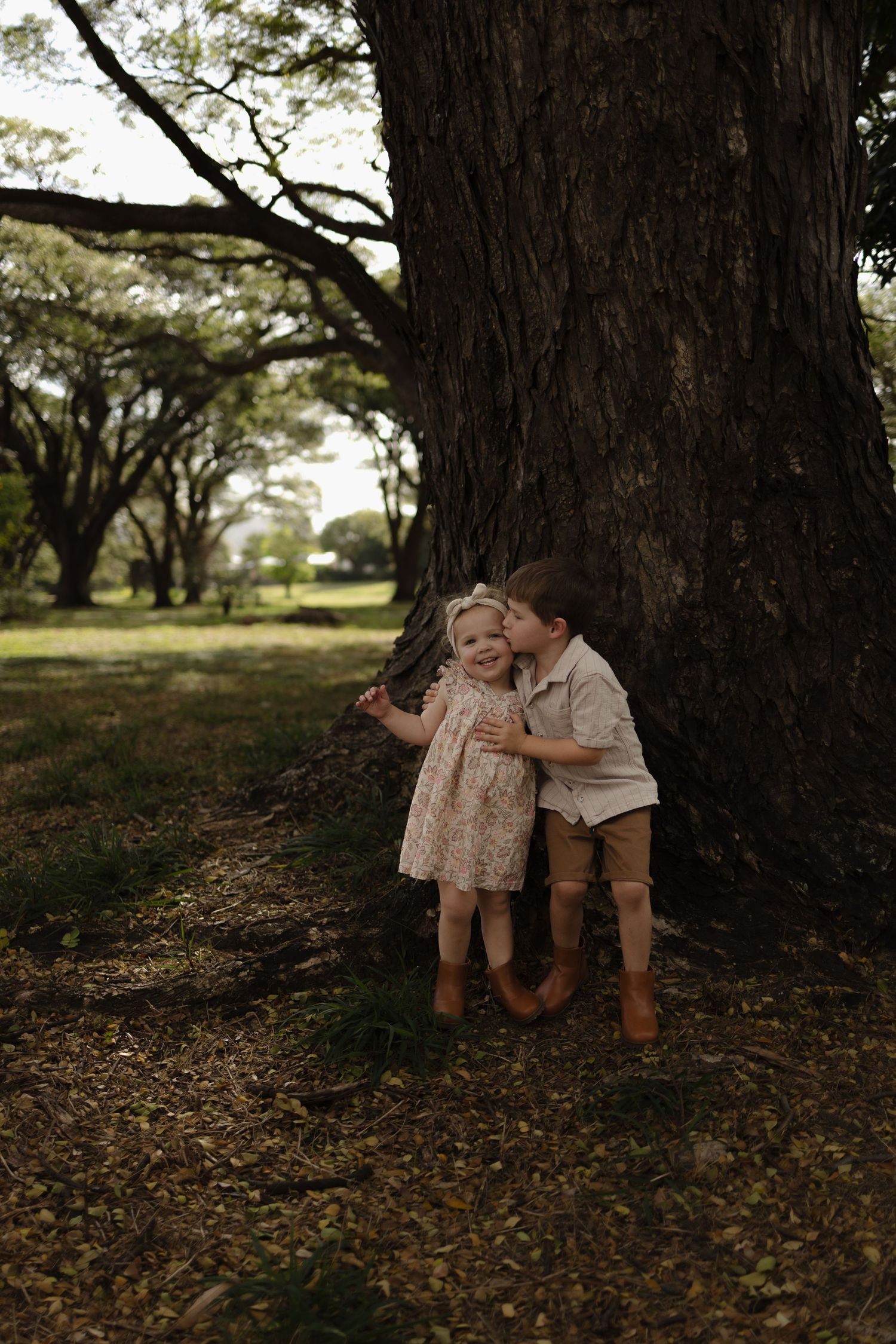 Two children rest together against a large tree trunk in dappled sunlight.