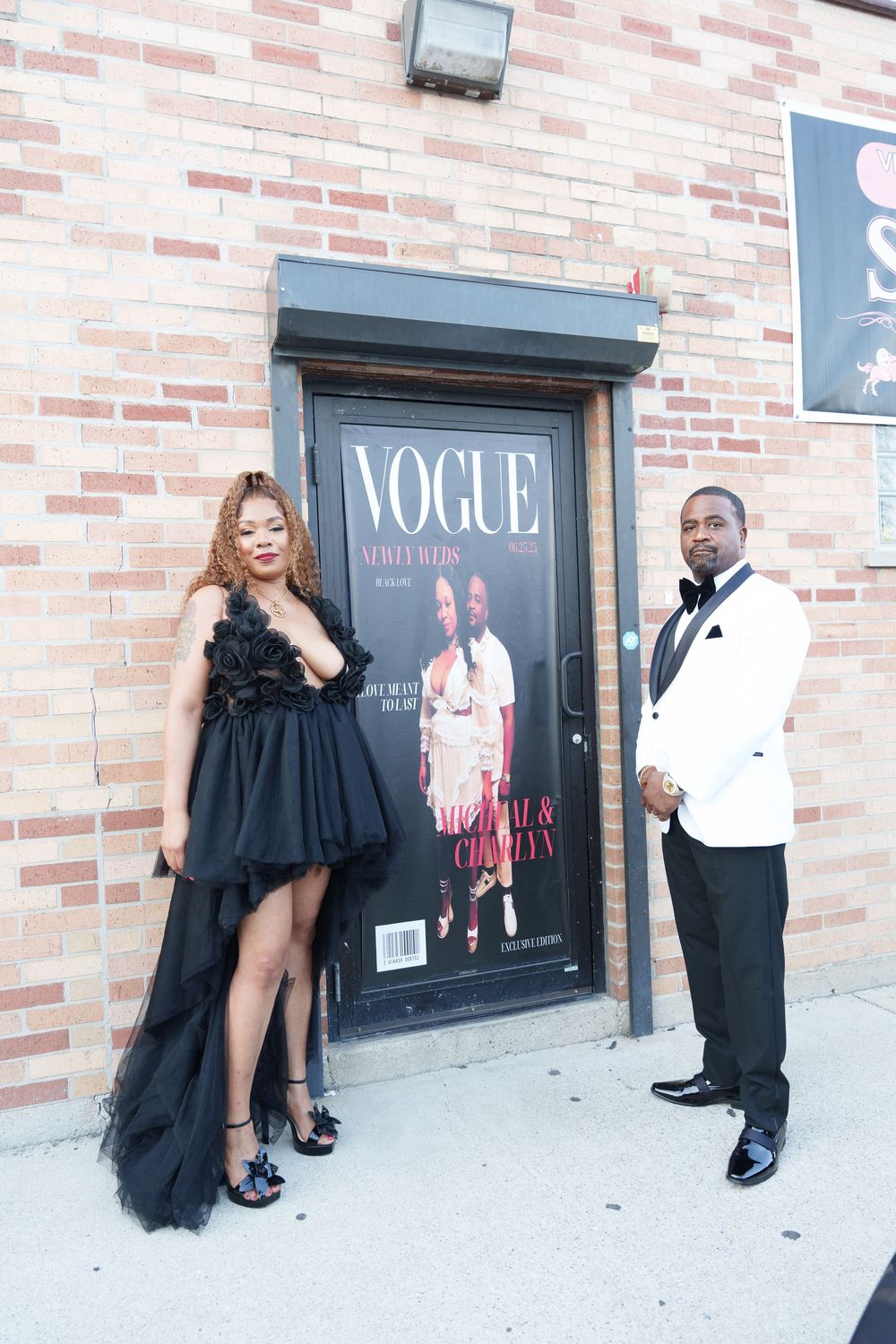 A couple poses in formal attire in front of a Vogue magazine poster on a brick wall.