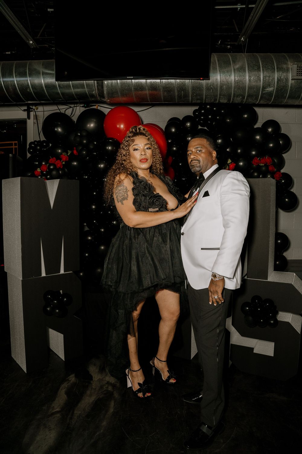 A couple enjoys dancing and posing together at a party decorated with black and red balloons in a dimly lit room.