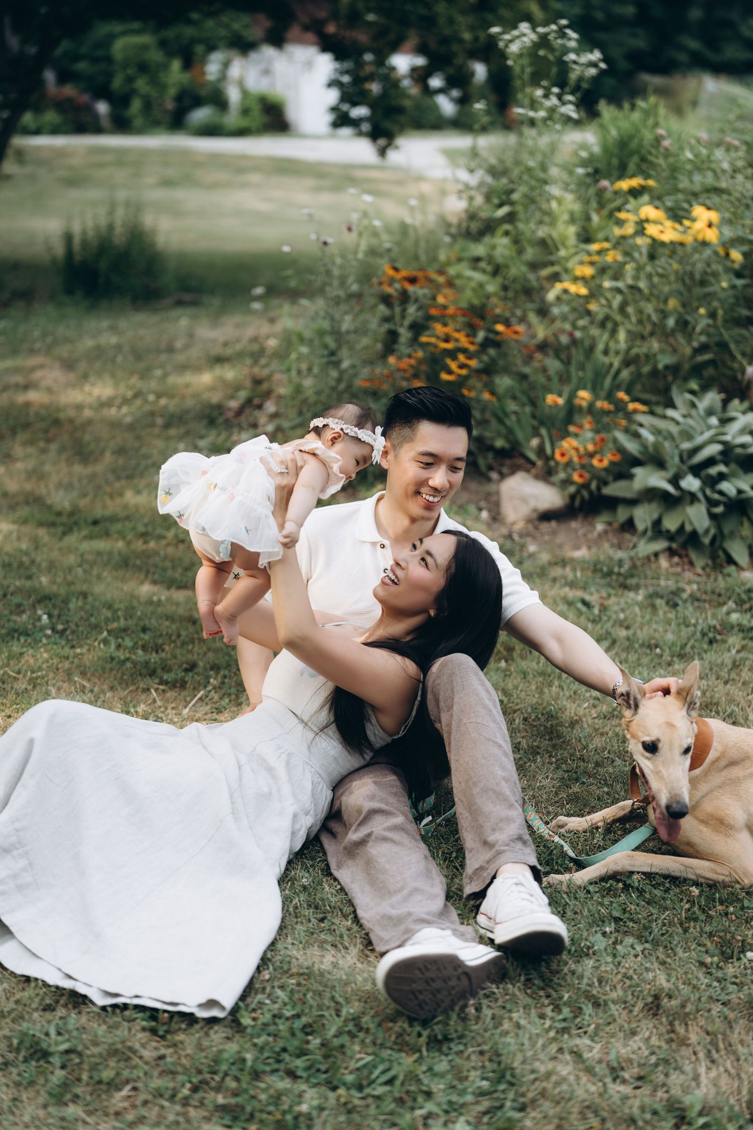 A couple relaxes on grass with their baby and dog beside orange flowers in a garden setting.