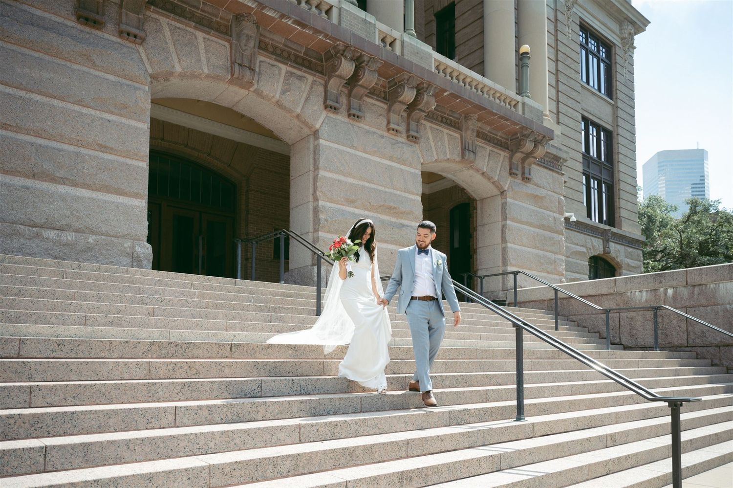 A bride in a flowing white dress and veil stands with a suited person in a covered brick colonnade walkway.