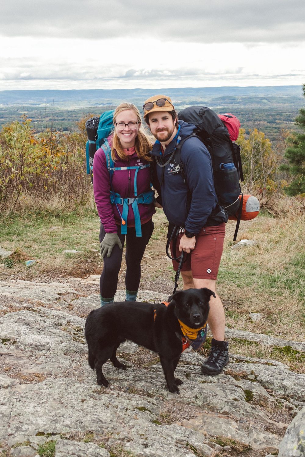 Hikers and their black dog pose together on a rocky mountain overlook.