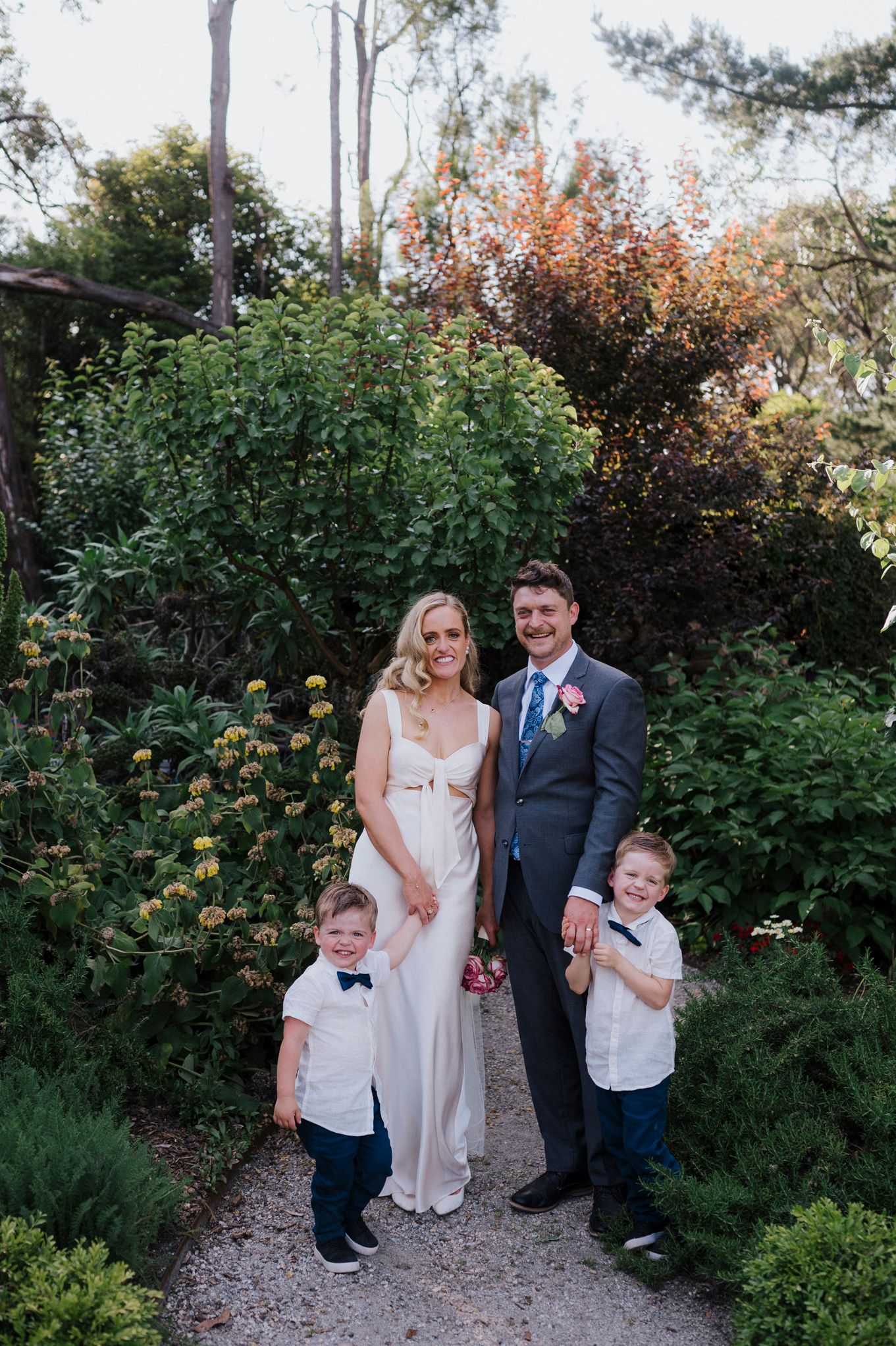 A family poses together on a garden path lined with lush greenery and autumn foliage in the background.
