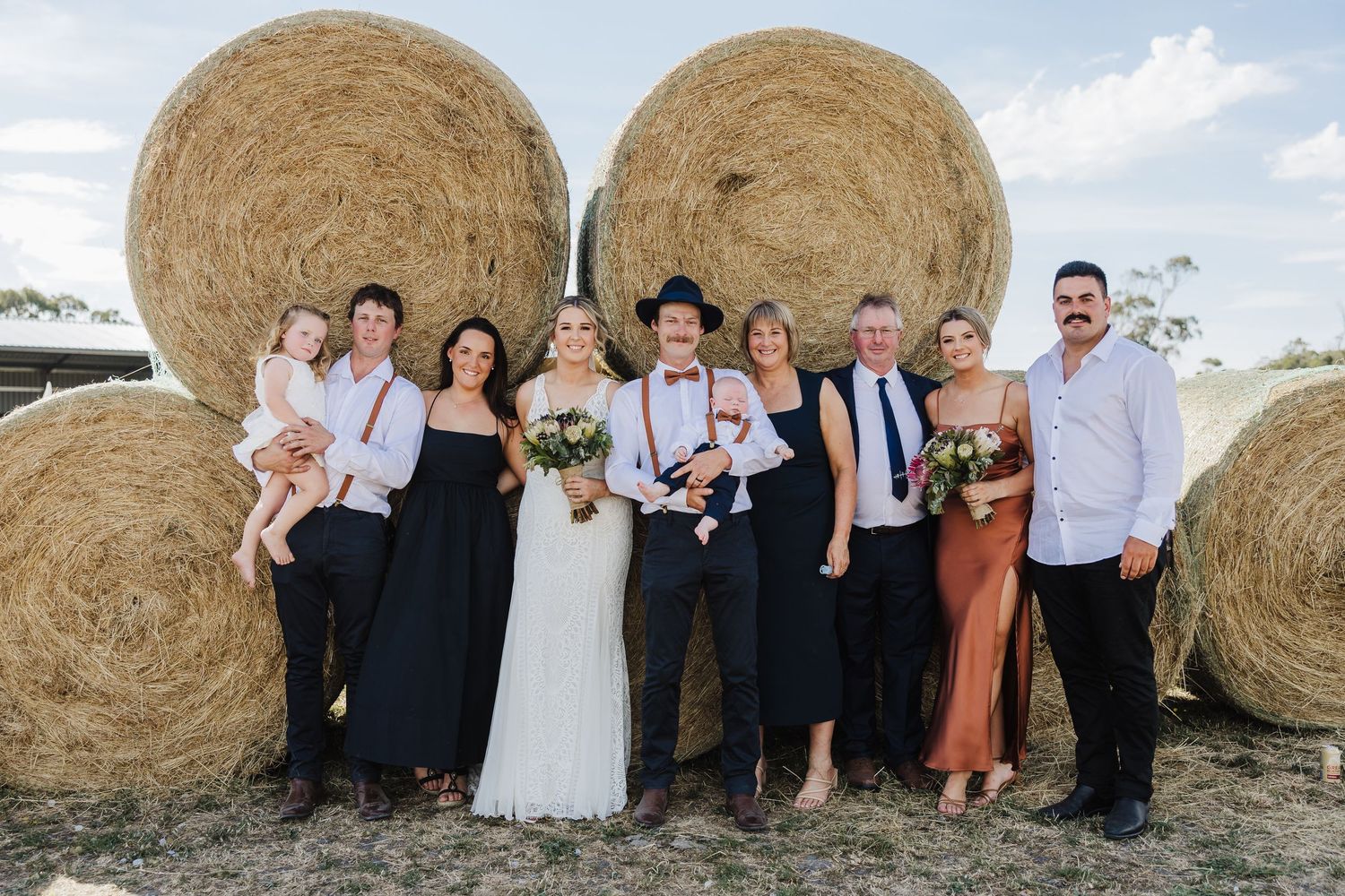 Wedding party members pose together for photos next to large round hay bales in a rustic outdoor setting.