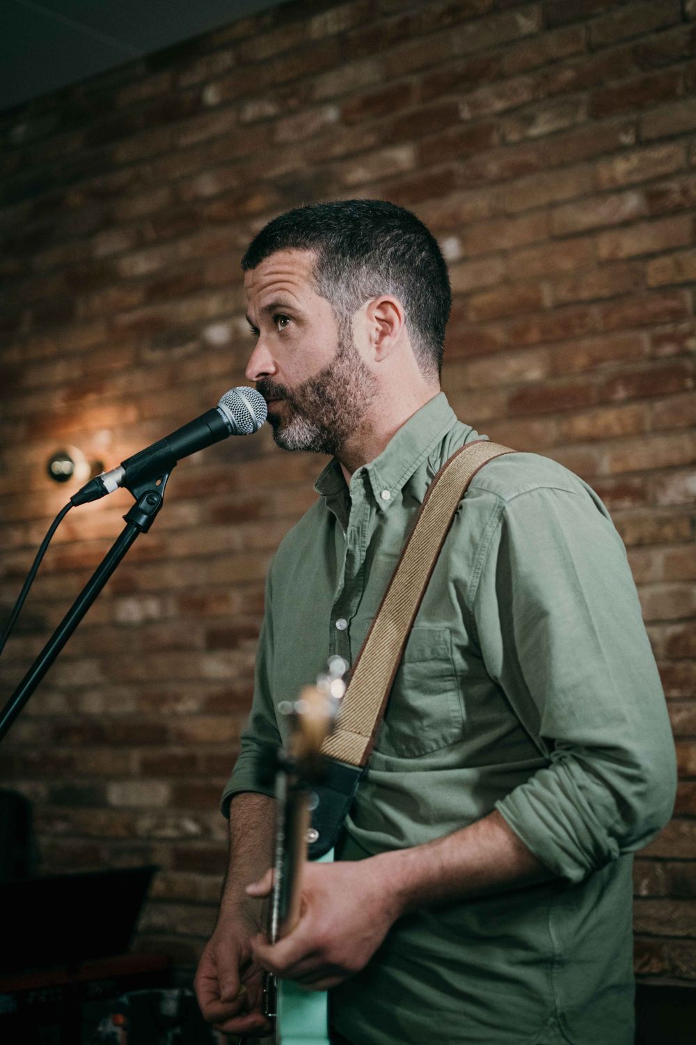Performer with guitar singing into microphone against brick wall backdrop.