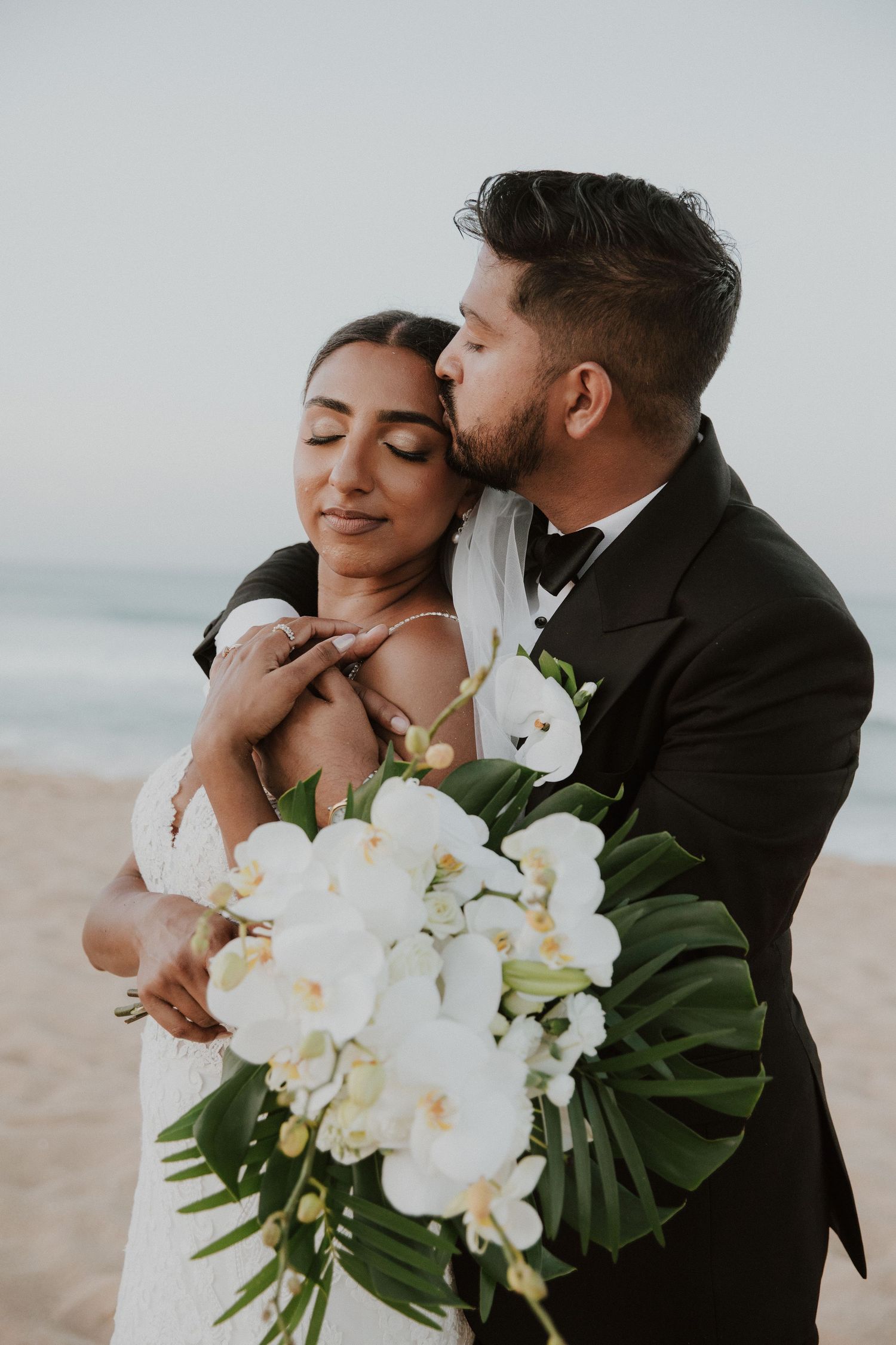 Intimate beach wedding moment with tropical white orchid bouquet and monstera leaves.