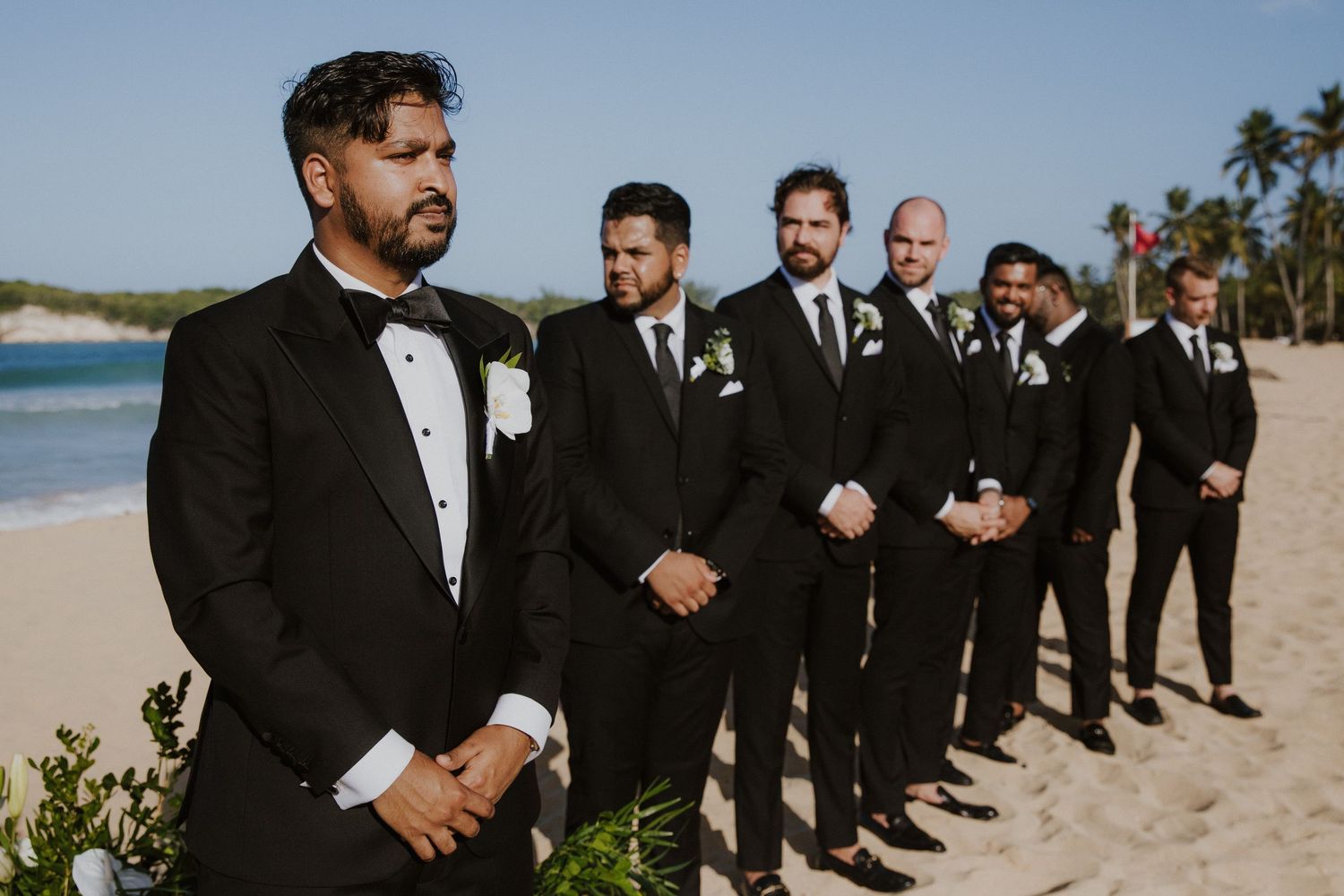 Groomsmen lined up on beach wearing black tuxedos with ocean backdrop.