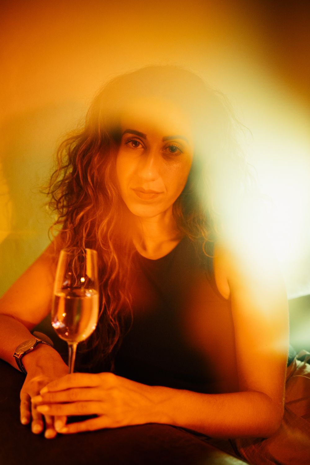 Woman with curly hair holding a wine glass in warm golden ambient light at a social gathering.