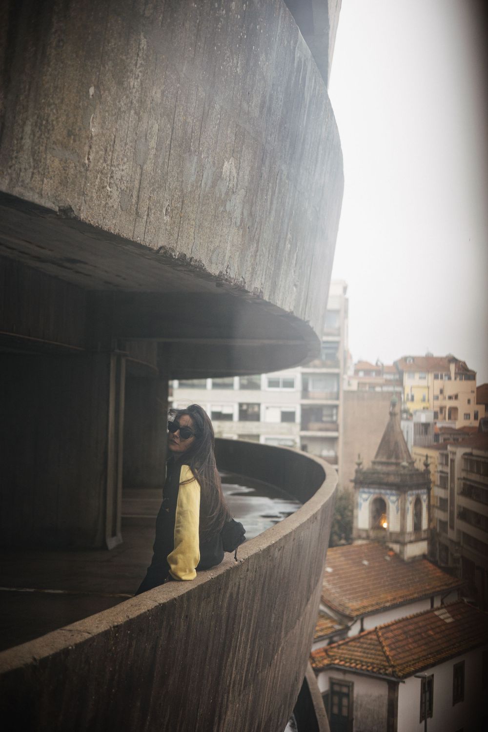 Woman in sunglasses poses on a curved concrete balcony overlooking a European cityscape with historic buildings.
