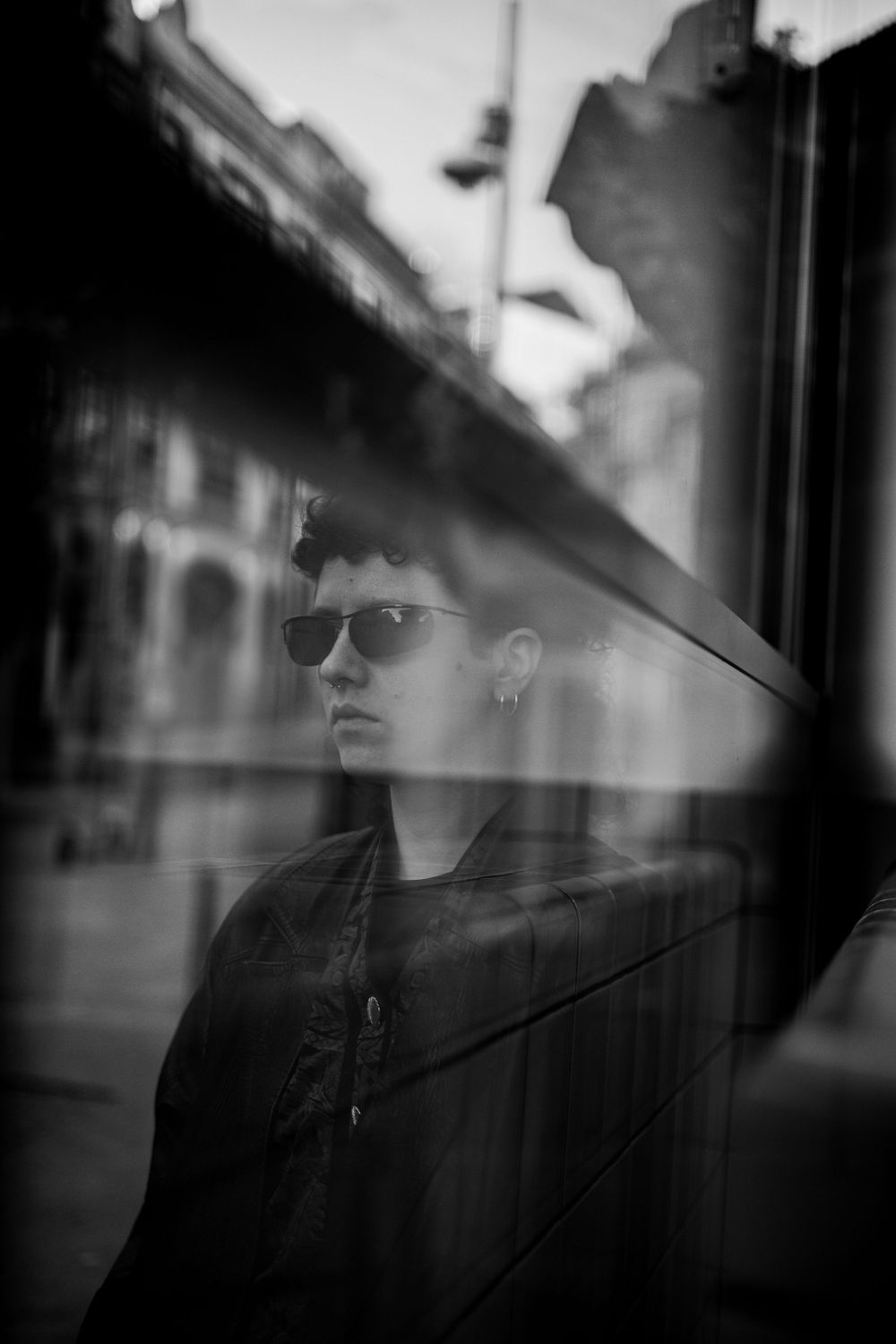Black and white portrait of a person wearing sunglasses reflected in a city window.