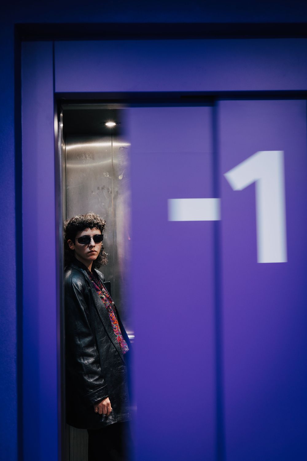 Young man in leather jacket and sunglasses standing in elevator doorway with purple wall showing -1 floor sign.