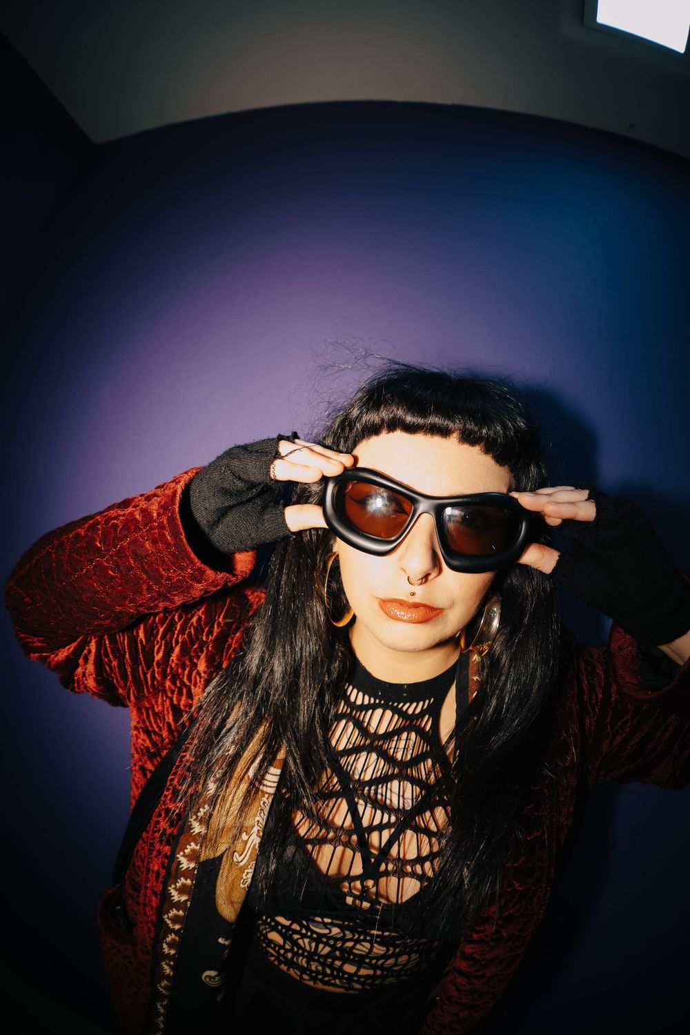 Woman in red jacket and fishnet top adjusts dark sunglasses against purple background.