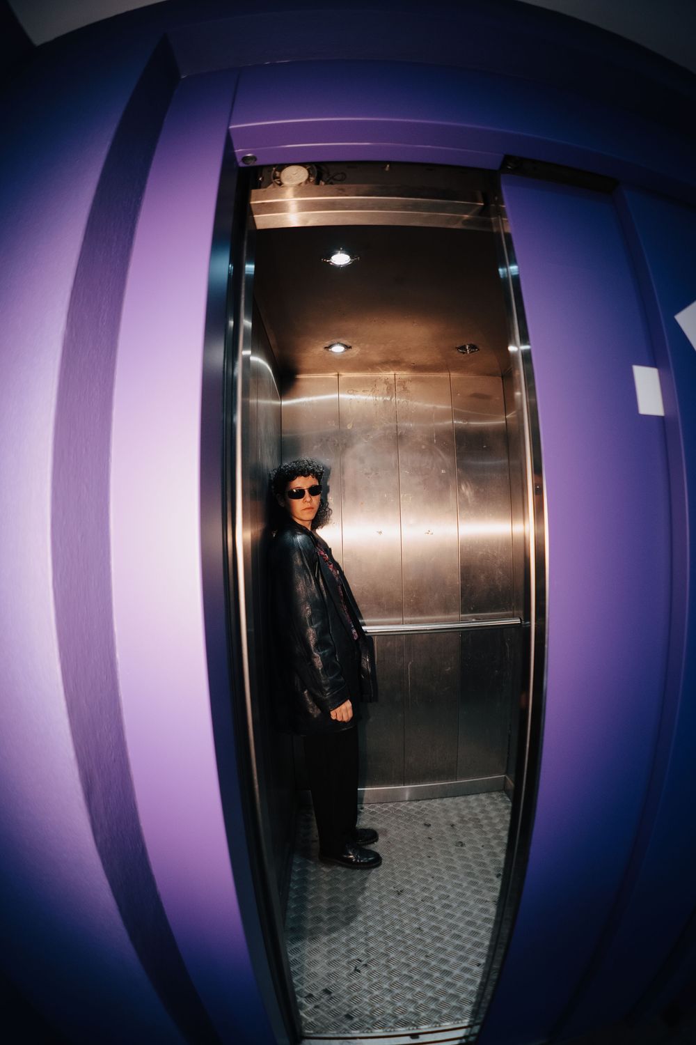 Person in black leather jacket and sunglasses standing inside a metallic elevator with purple walls.