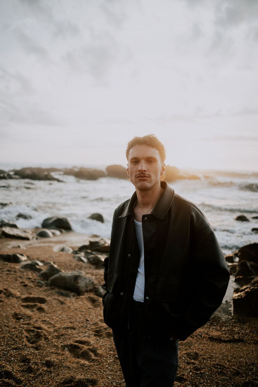 Young man in dark jacket standing on rocky beach at sunset with ocean waves in background.
