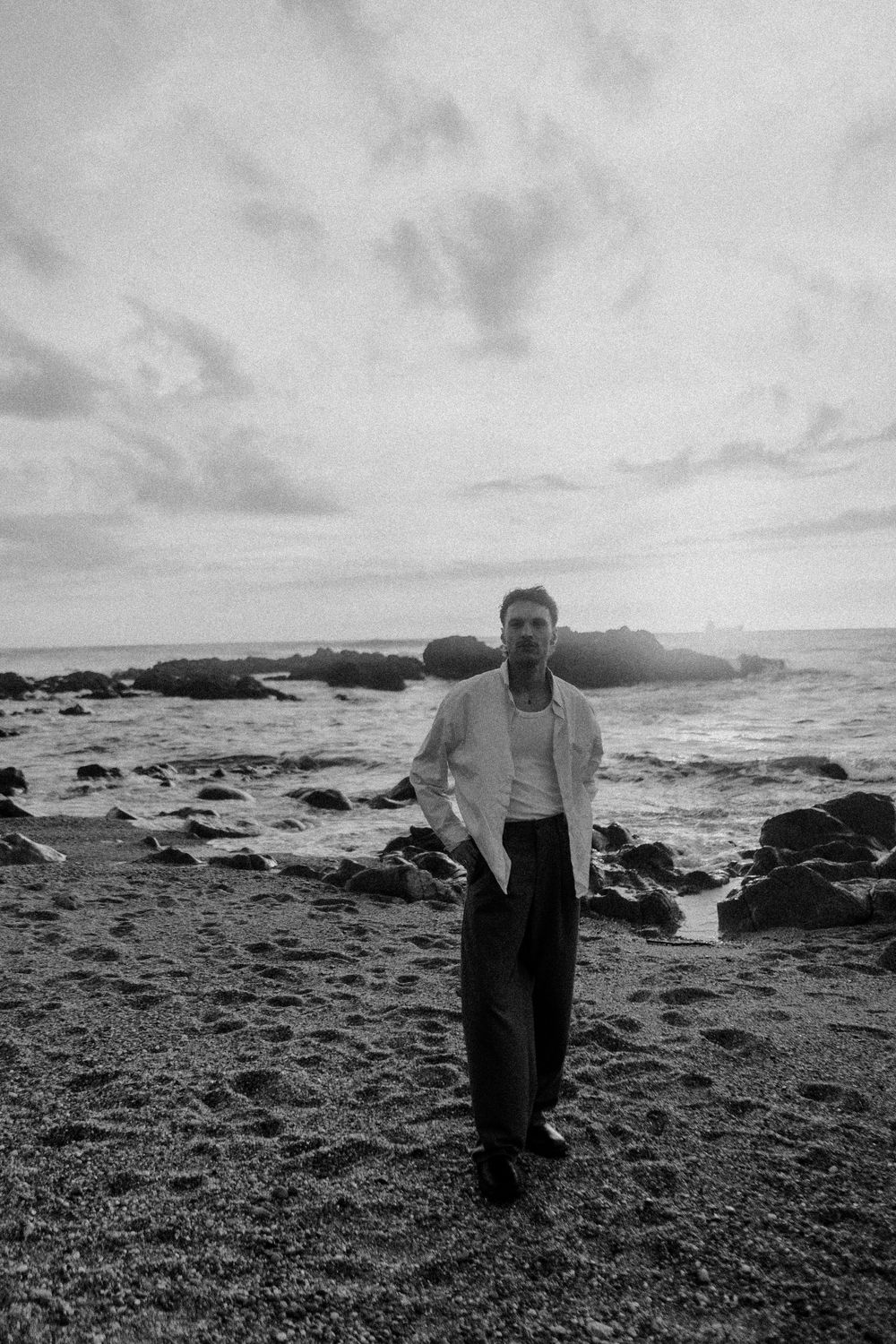 Black and white photo of a man in a white blazer standing on a rocky beach with dramatic cloudy skies.