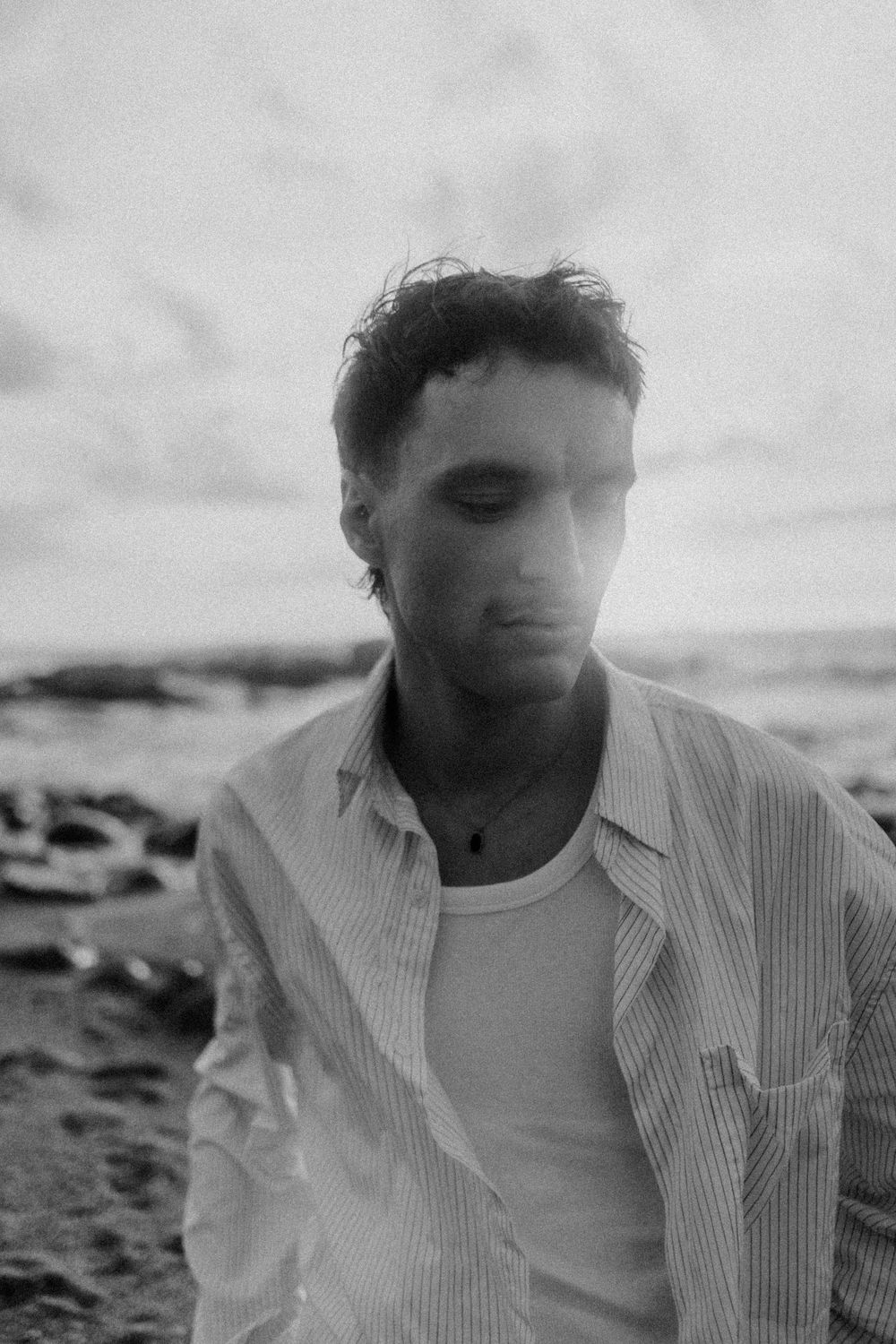 Black and white portrait of a young man in a striped shirt standing by the ocean under a cloudy sky.