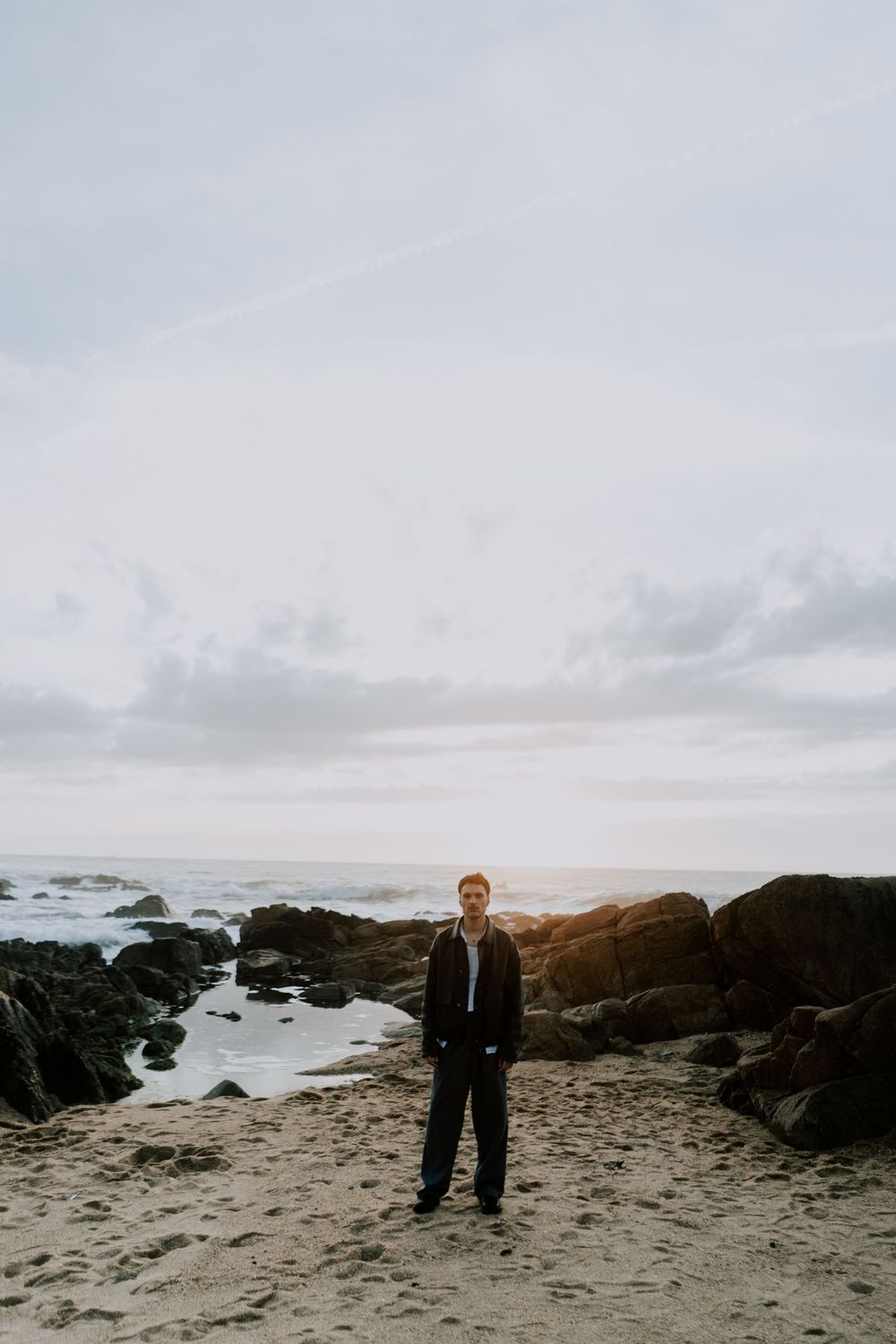 A man in a dark jacket stands on a rocky beach with ocean waves and cloudy sky in the background.