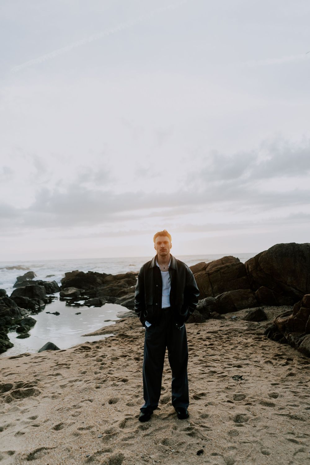 A man in a dark jacket and black pants stands on a sandy beach with rocky coastline and cloudy sky behind him.