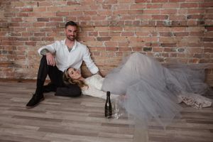 Wedding couple relaxing on wooden floor with champagne bottle by brick wall.
