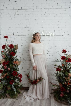 Bridal portrait with two-piece wedding dress featuring lace top and tulle skirt against white brick wall with red floral arrangements.