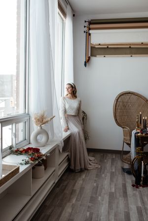 A bride in a lace top and tulle skirt stands by a window in a minimal room with peacock chair.