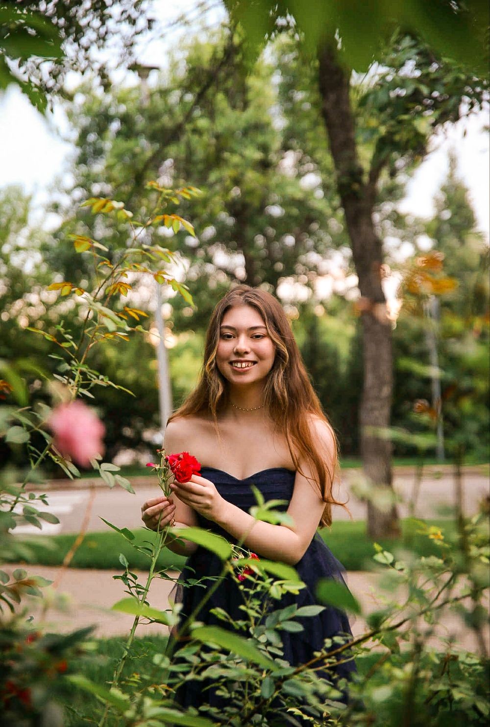 Smiling young woman in navy dress holding red roses in a lush garden setting.