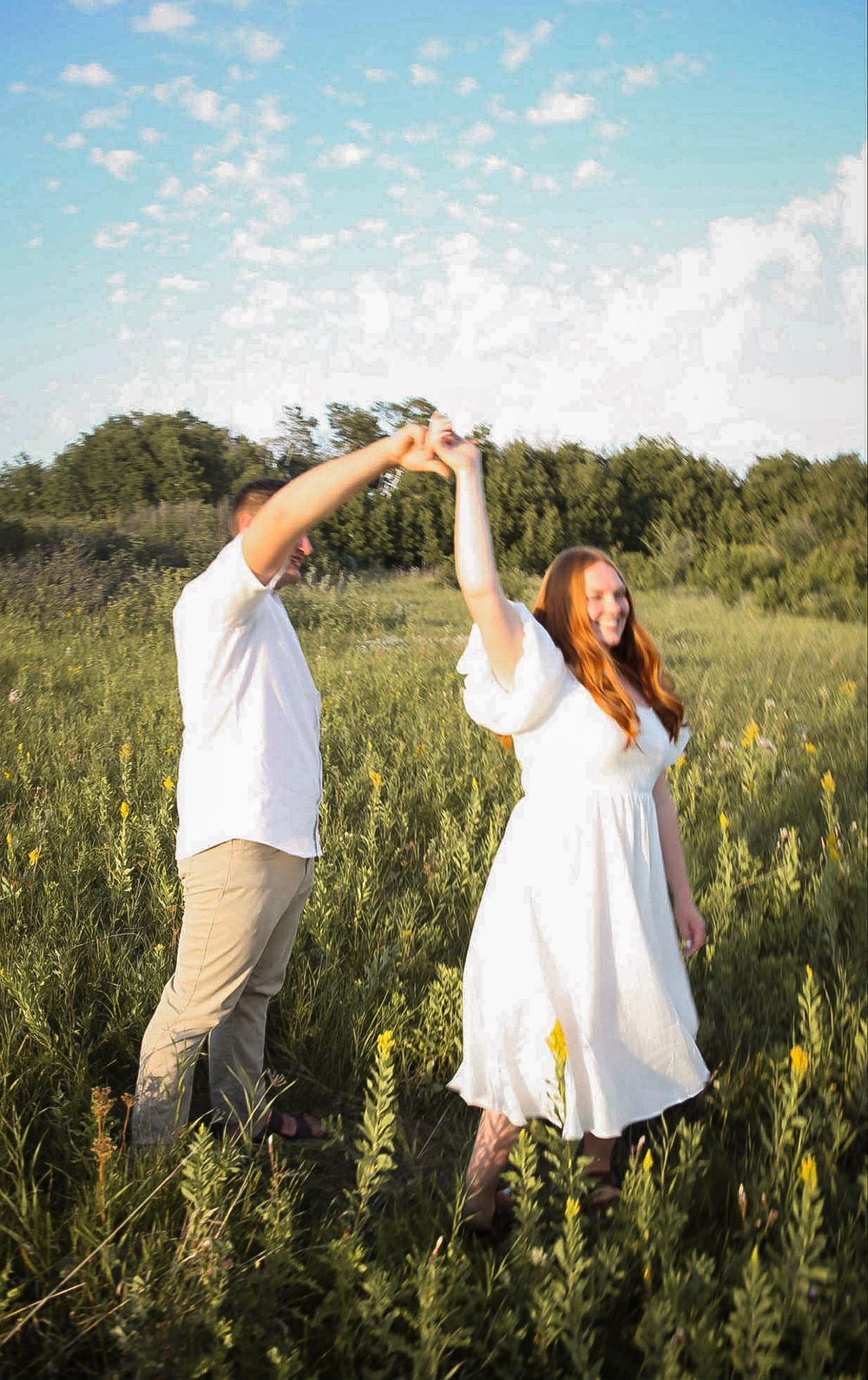 A couple dances joyfully in a sunny green meadow, the woman in a white dress twirling as they hold hands under a blue sky.