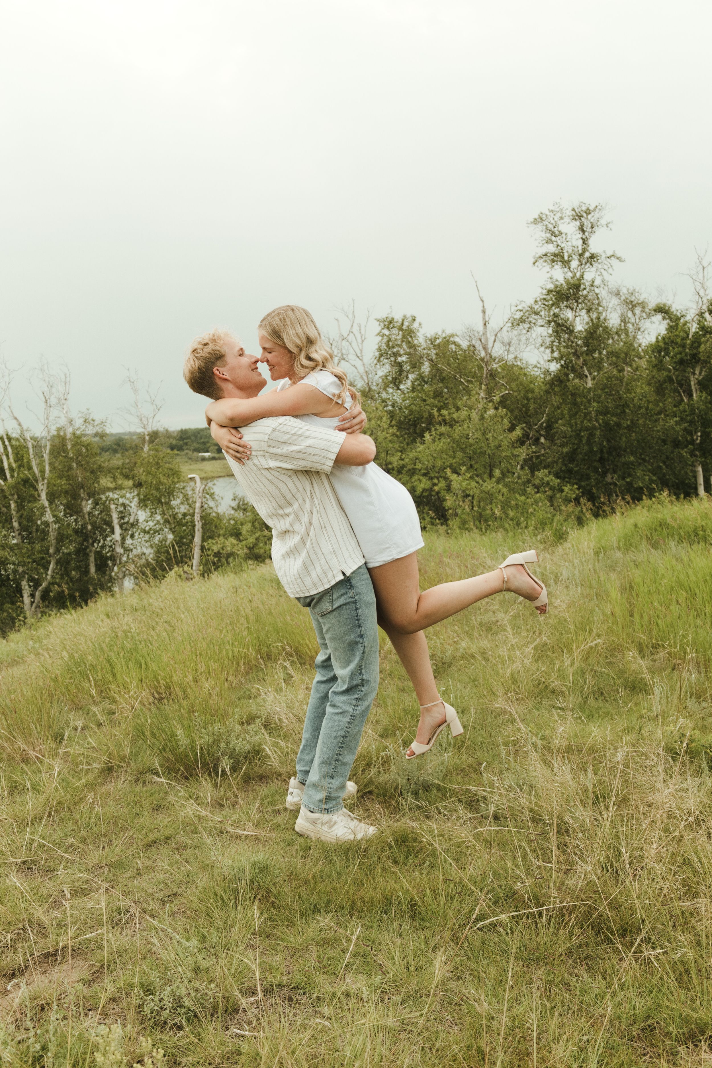 A couple embraces and kisses outdoors on a grassy hill, the man lifting the woman in a white dress.