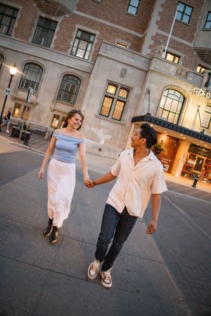 A couple holds hands and walks together on a city street, smiling, with a historic brick building in the background.