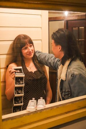Pregnant woman holding ultrasound photos smiles at partner in denim jacket, with baby shoes displayed on windowsill.