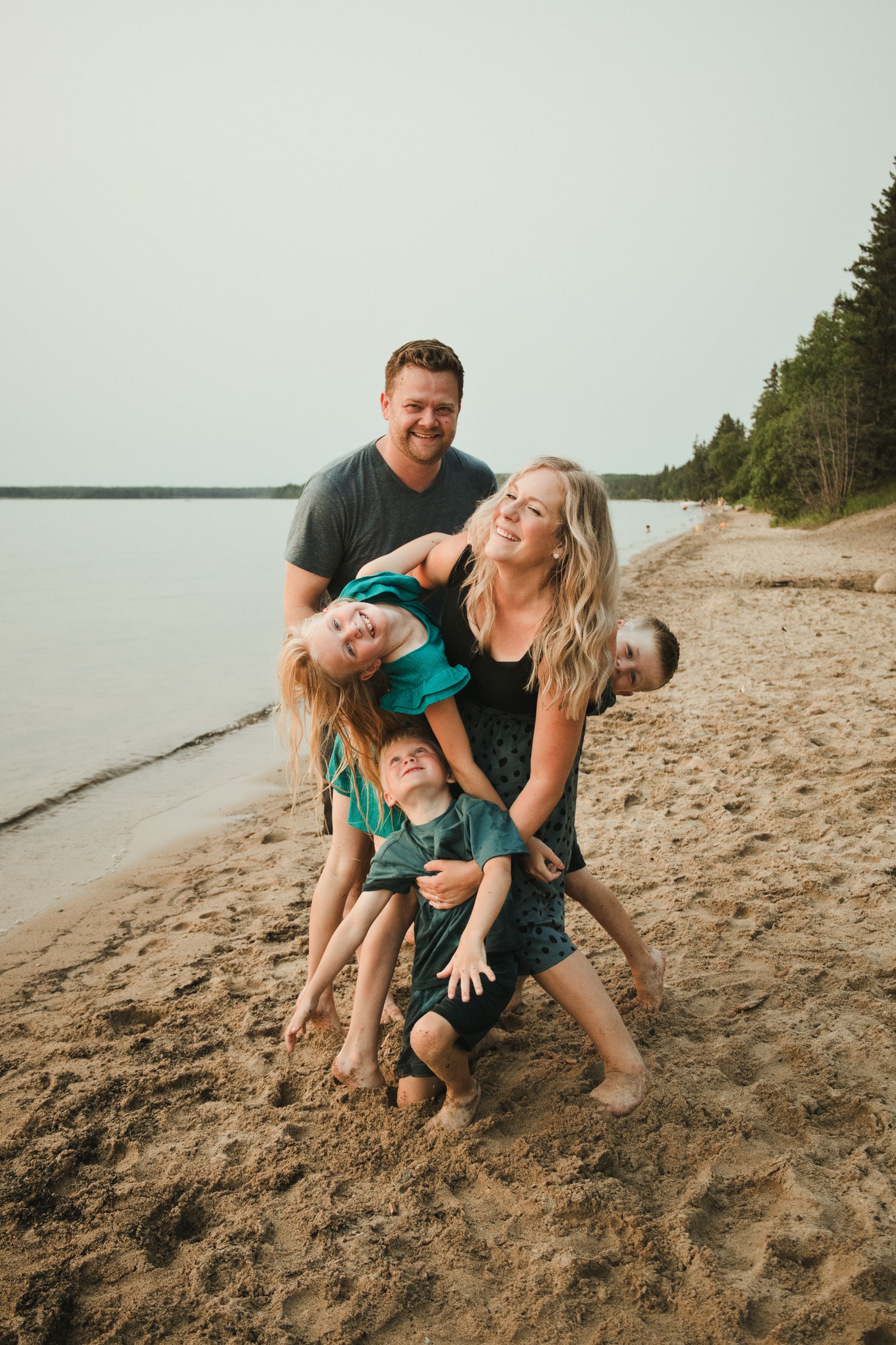 Happy family of four playing on a sandy beach near a lake, with tall pine trees in the background.