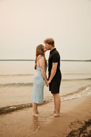 A couple kissing on a sandy beach shore, woman in light blue dress and man in black outfit, standing barefoot in shallow water.