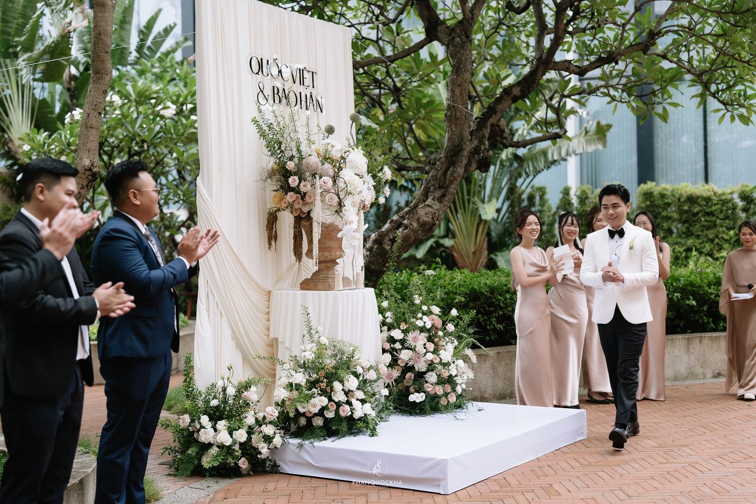Groom in white tuxedo walking toward floral backdrop as guests applaud at outdoor wedding ceremony.
