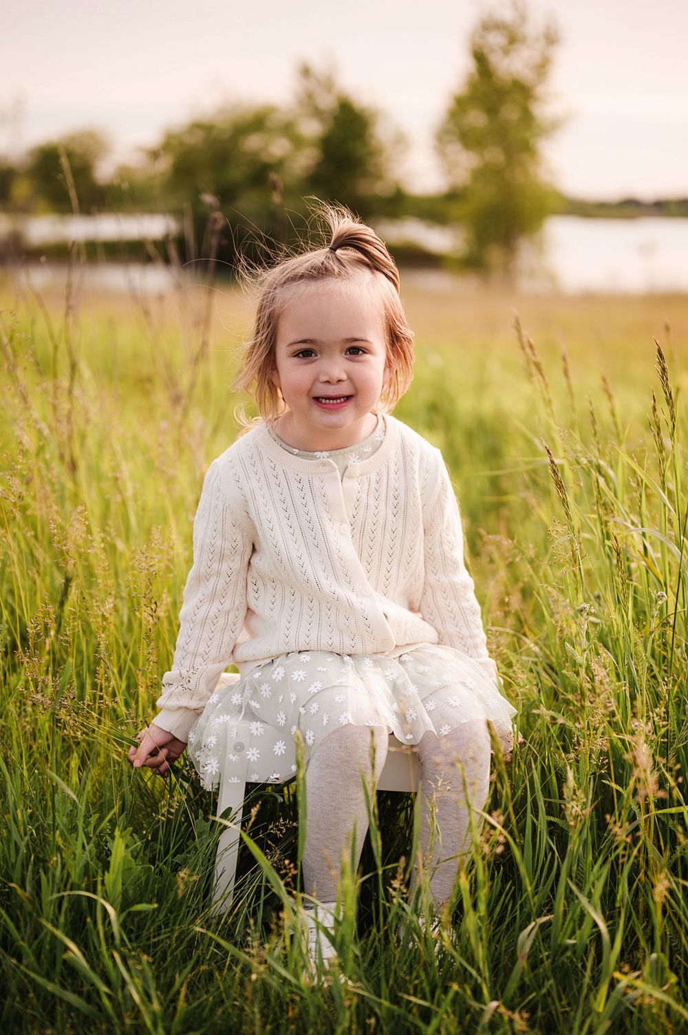 A child wearing a white sweater and tights sits joyfully in tall green grass during a sunny outdoor photo session.