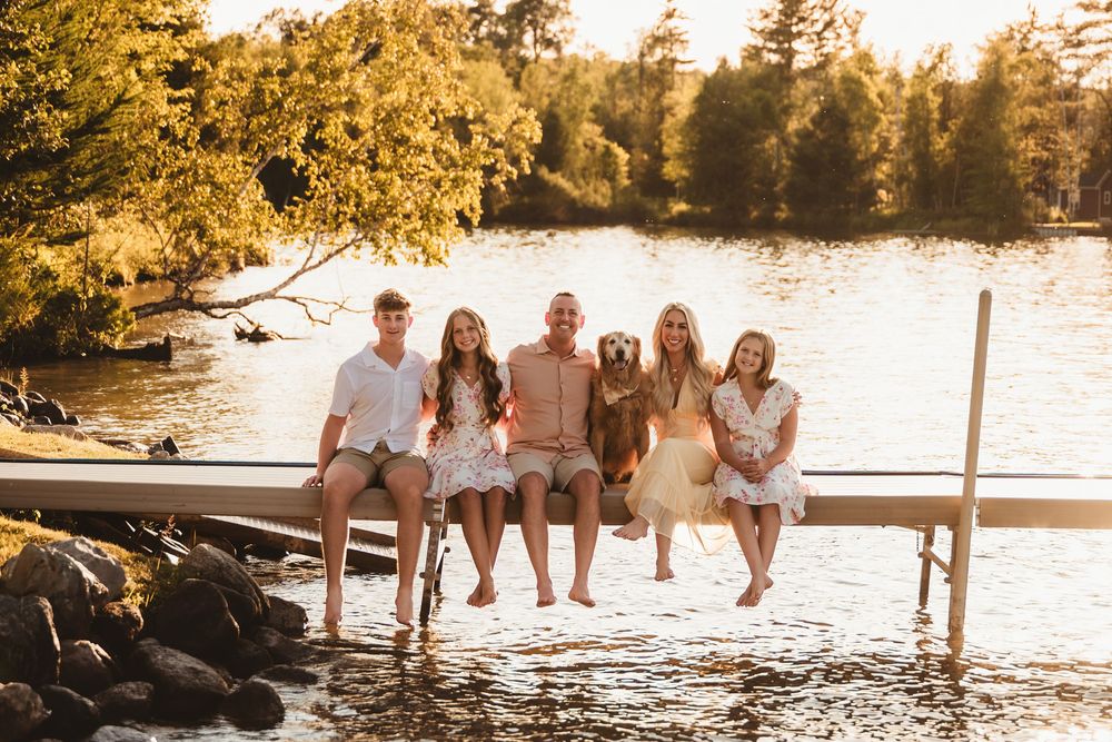 Group sits together on wooden dock during golden sunset at lake surrounded by pine trees.