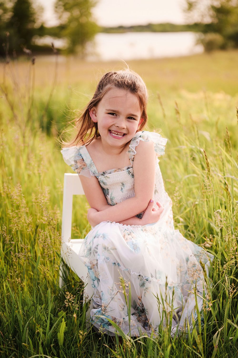A child in a floral white dress sits on a chair in a sunlit meadow during golden hour.