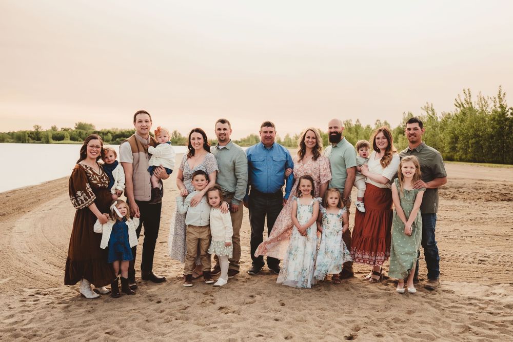 Large family group poses together on a sandy beach with a lake and greenery in the background at sunset.