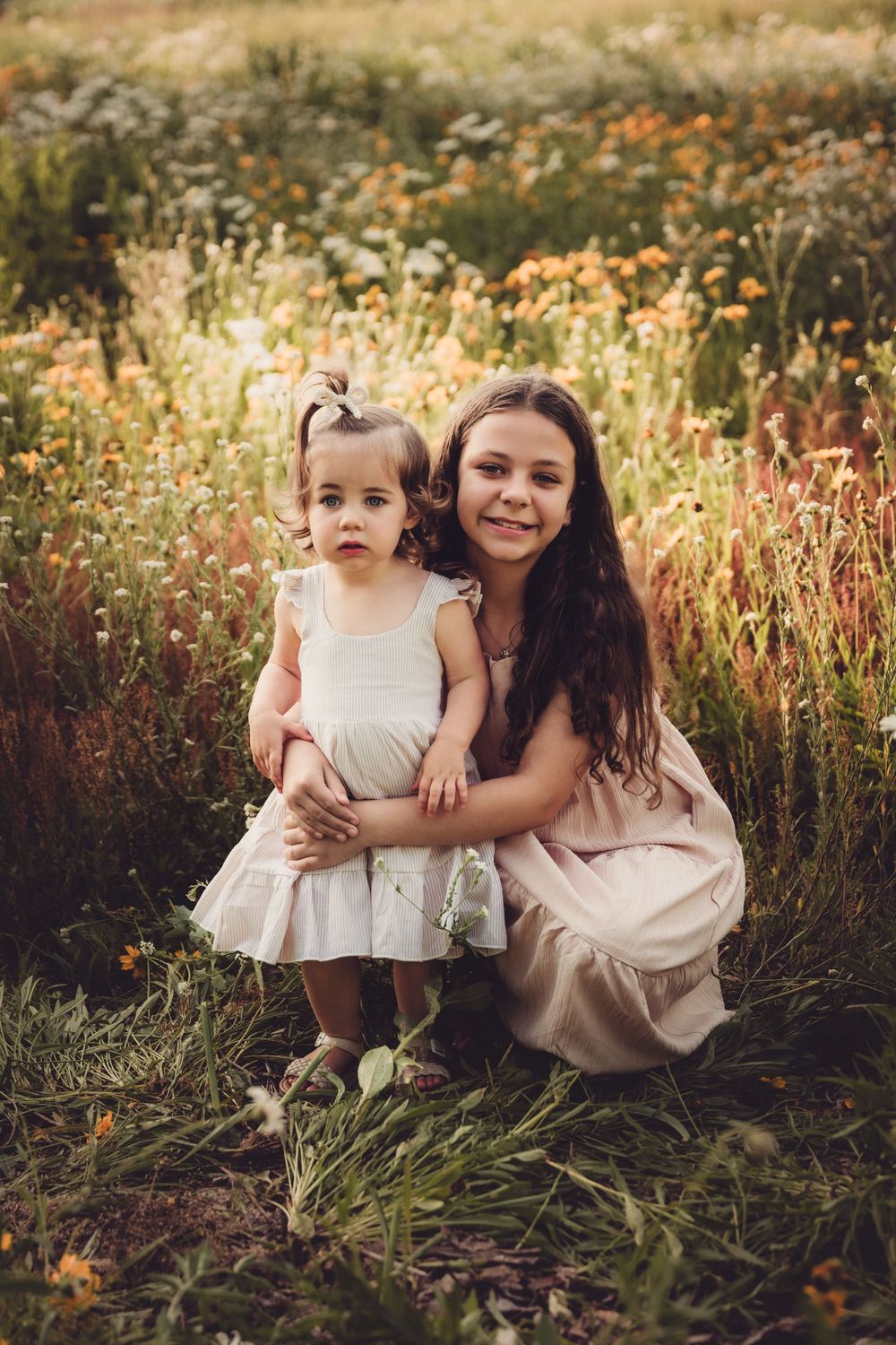 Two people in light dresses share a tender moment among wildflowers in a dreamy sunset meadow.