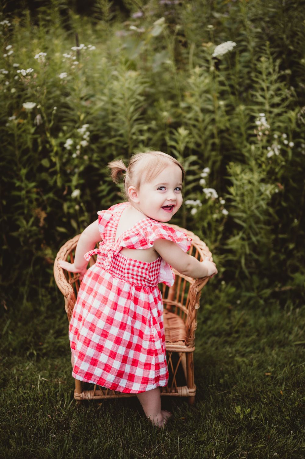 A red and white gingham dress adorns a small child sitting in a wooden chair outdoors among wildflowers and greenery.