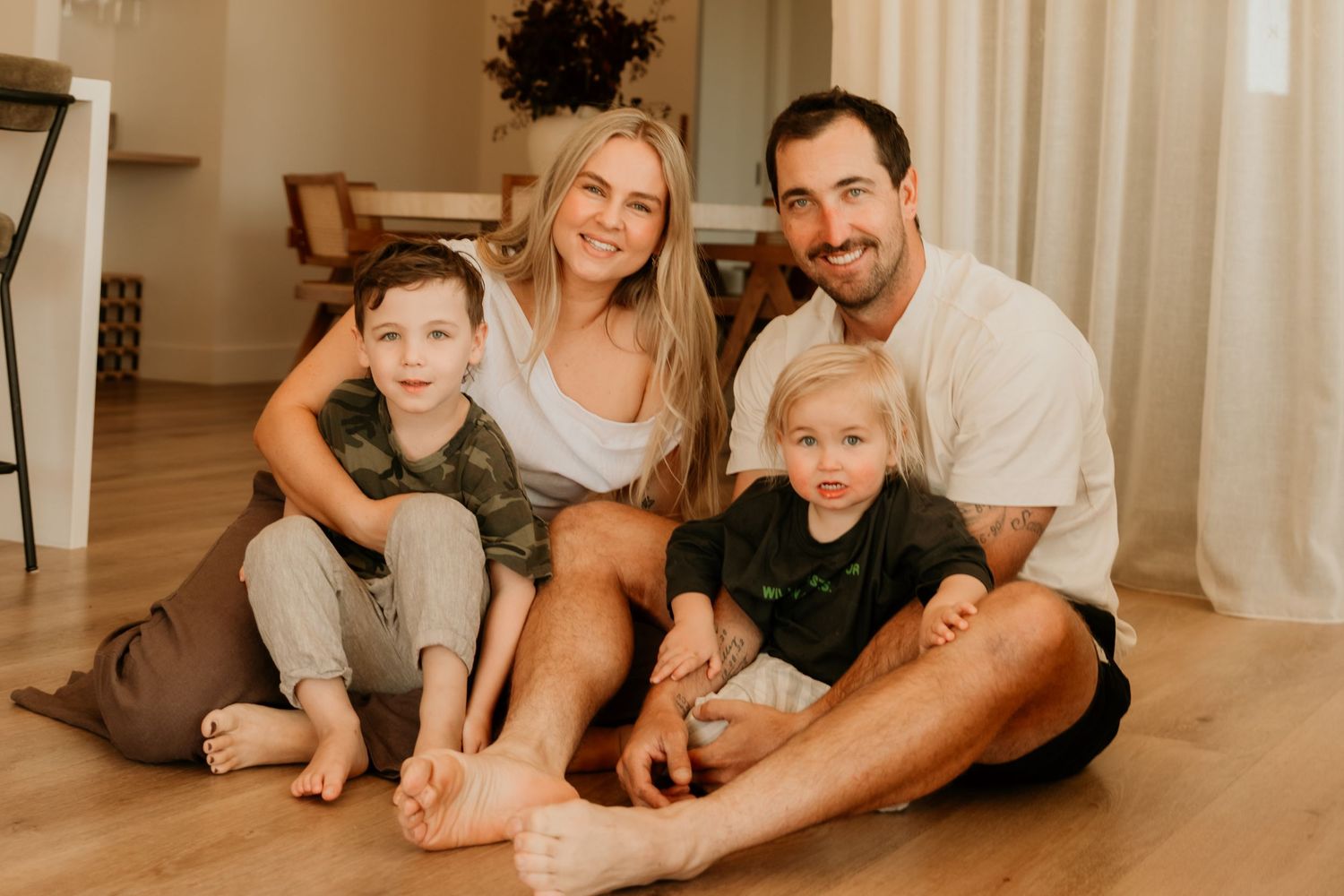 Happy family of four sitting together barefoot on hardwood floor smiling at camera in modern home.